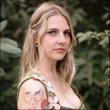 A person with blonde hair and tattoos
Description automatically generated